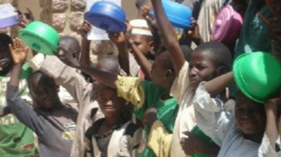 A crowd of Almajiri children in green and white caps, hands raised, gathered closely together outdoors.