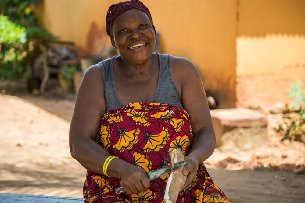 A woman sitting outside a house, smiling, peeling a cassava root with a knife.