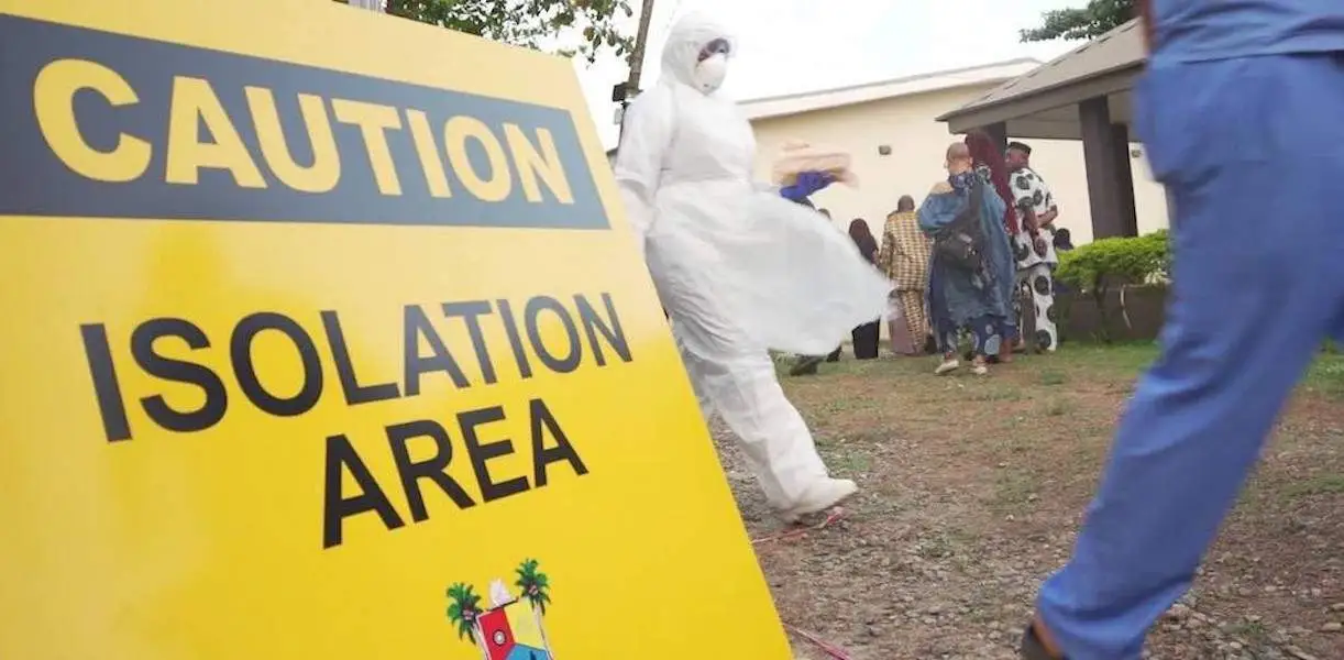A yellow “Caution — Isolation Area” sign in the foreground, with a health worker in full protective coveralls and waiting members of the public behind it.