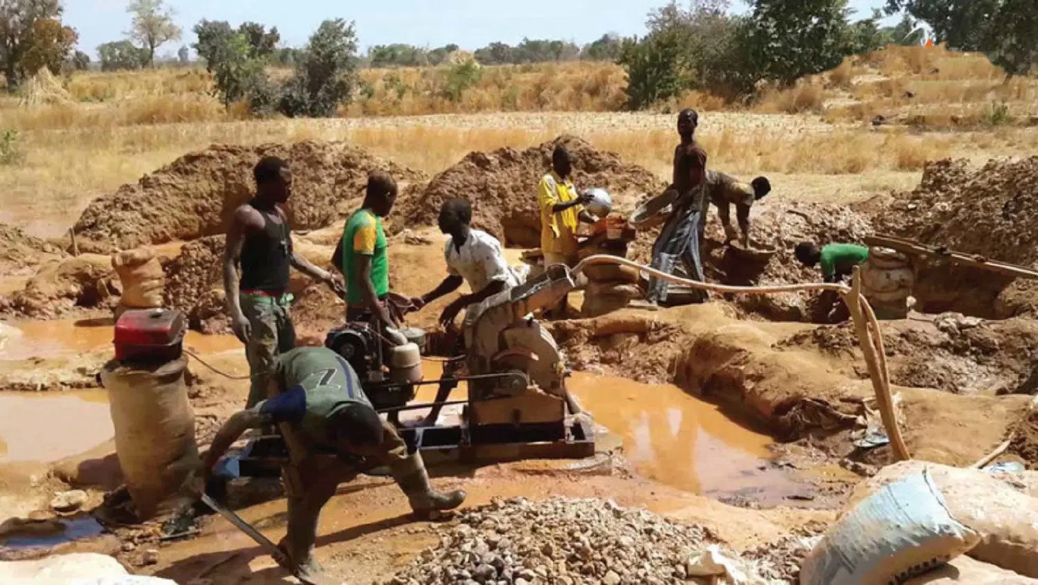 Artisanal miners working a flooded pit with a pump and sluice, sacks of ore stacked on the bank.