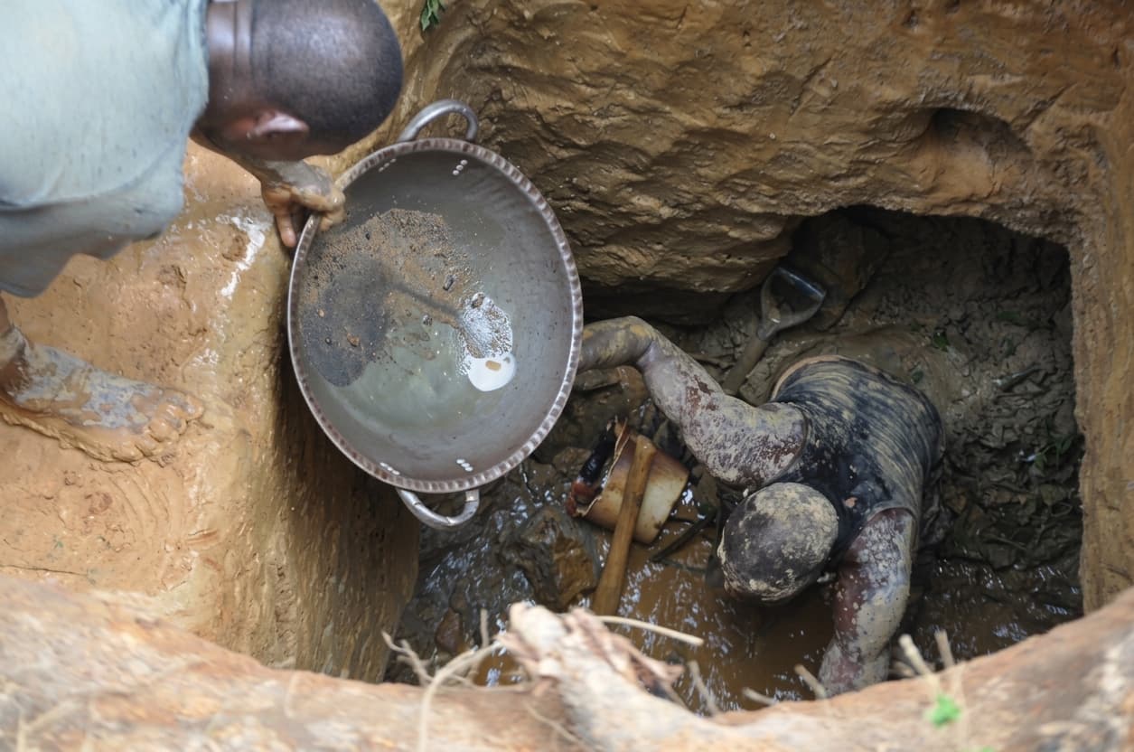 Two artisanal miners working a flooded pit by hand, one lowering a metal pan to the other below.
