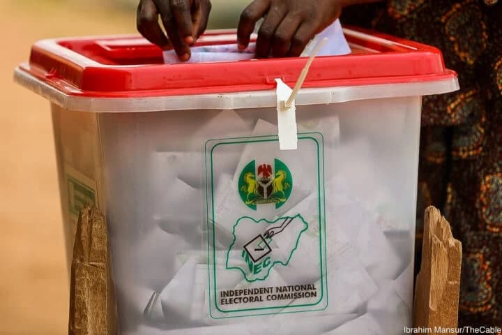 A voter pressing a folded ballot paper into the slot of a transparent INEC ballot box at a polling unit.