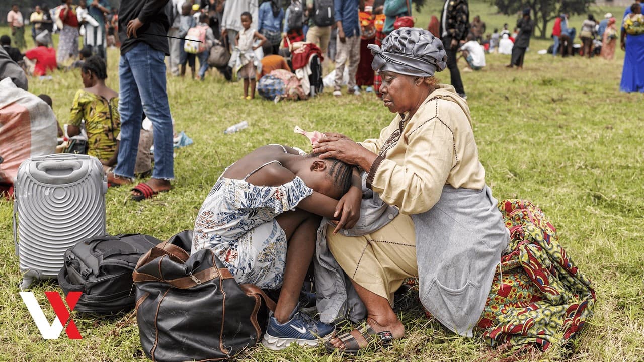 A woman comforting a child resting against her lap on open ground, with displaced families and their belongings gathered behind them.