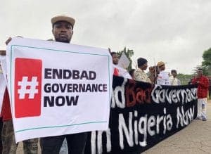 A protester holding a placard reading “#EndBad Governance Now” at the head of a march, with others carrying a banner behind him.