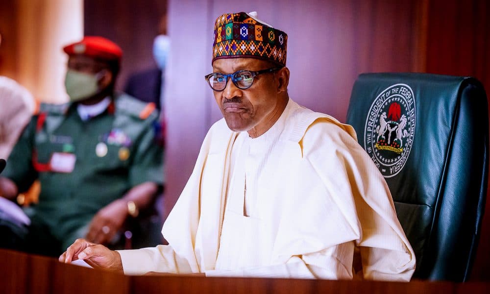 President Muhammadu Buhari seated at a desk beside the Nigerian flag, with a military aide standing behind him.