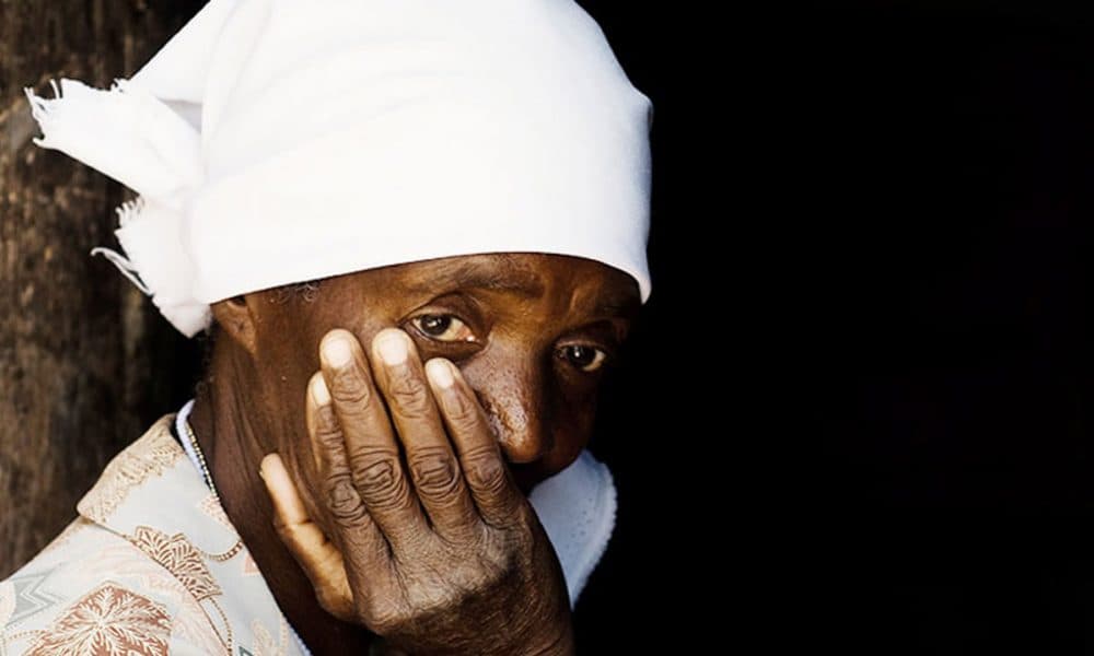 An elderly woman in a white head-tie resting her hand across her mouth, photographed against a dark background.