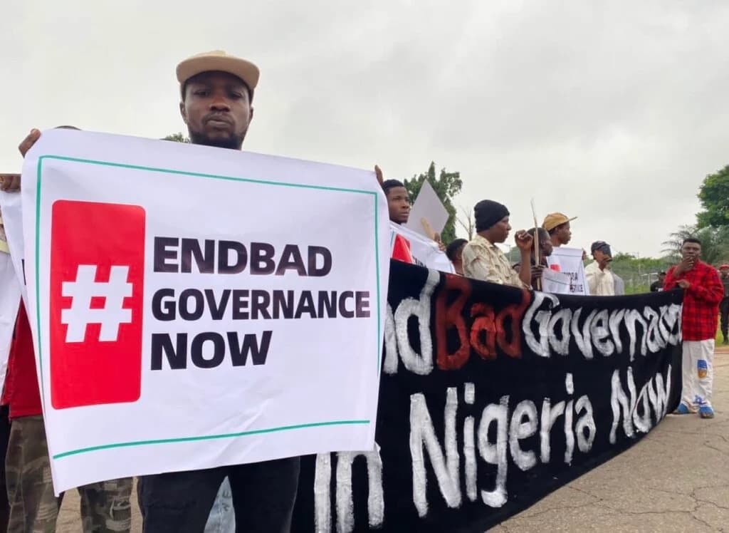 A protester holding a placard reading “#EndBad Governance Now” at the head of a march, with others carrying a banner behind him.