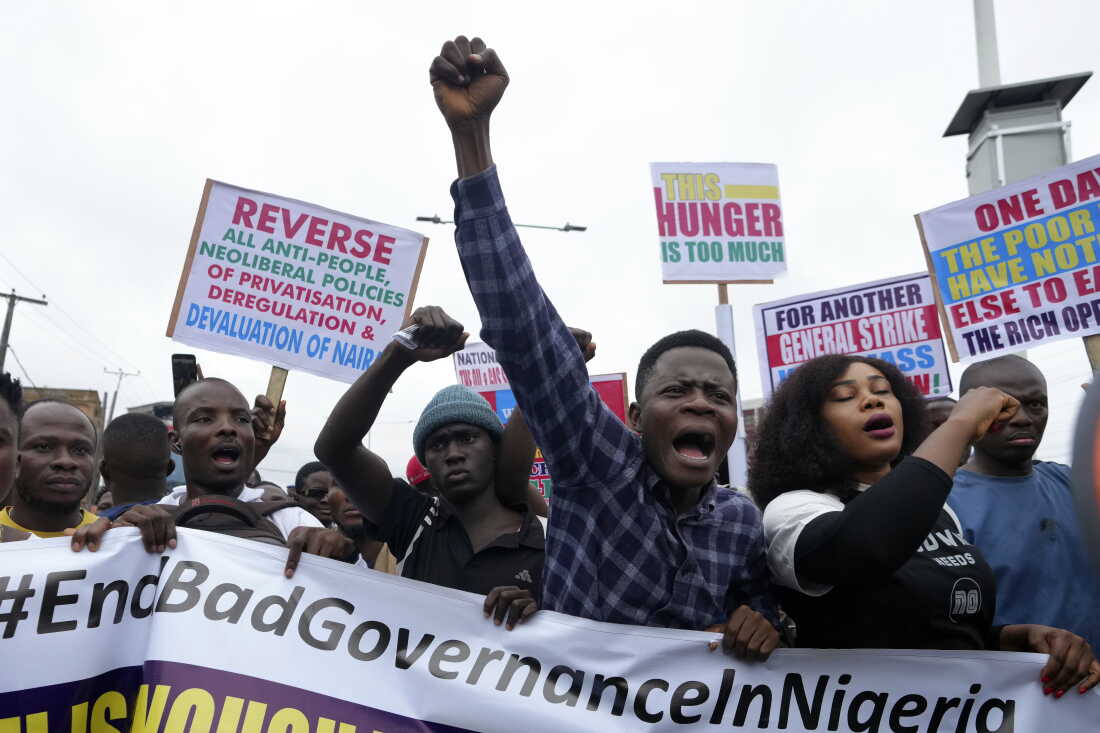 Protesters marching behind a banner reading #EndBadGovernanceInNigeria, one with a raised fist, others holding placards about the cost of living.